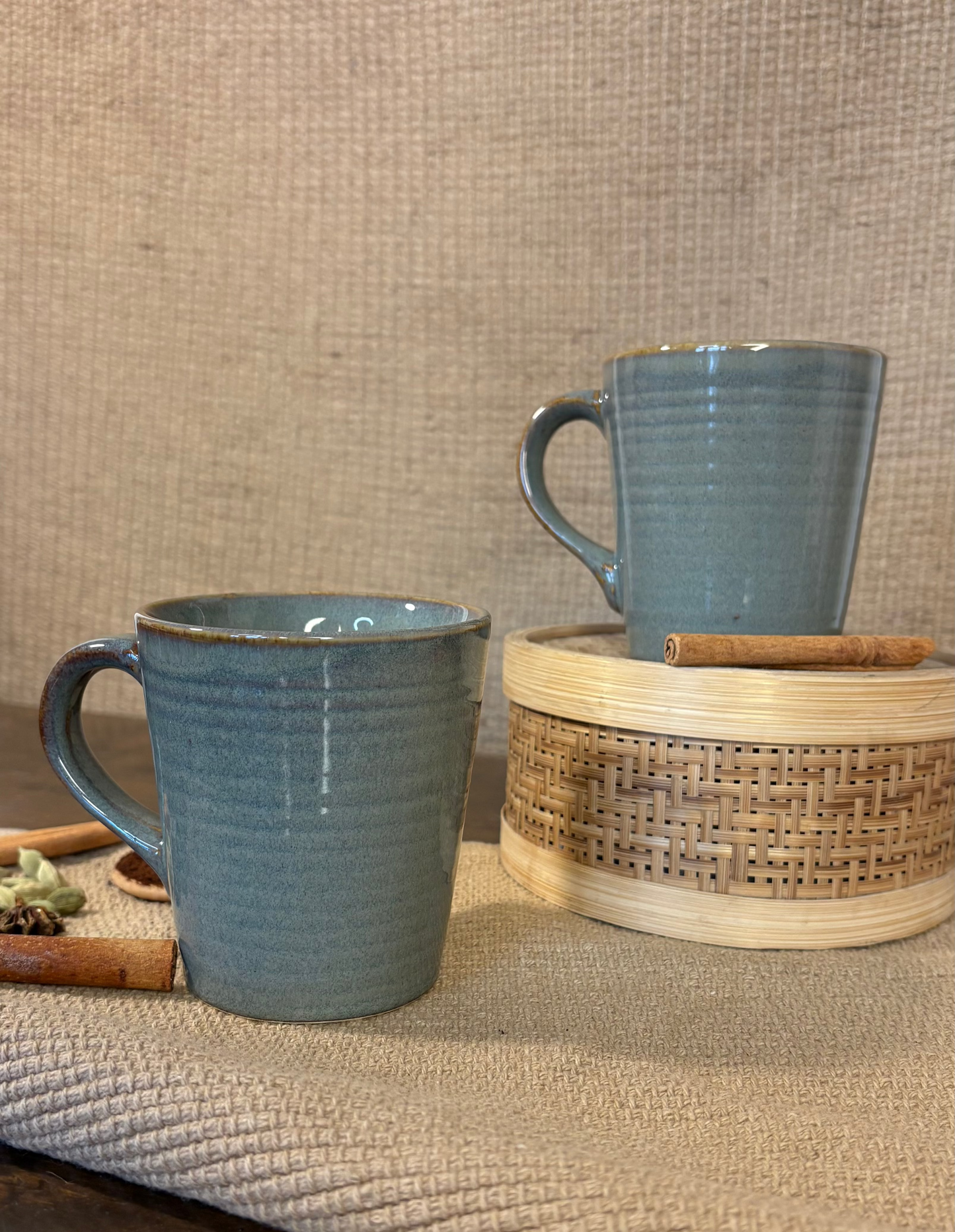 Two blue ceramic mugs on a textured surface with a woven basket.